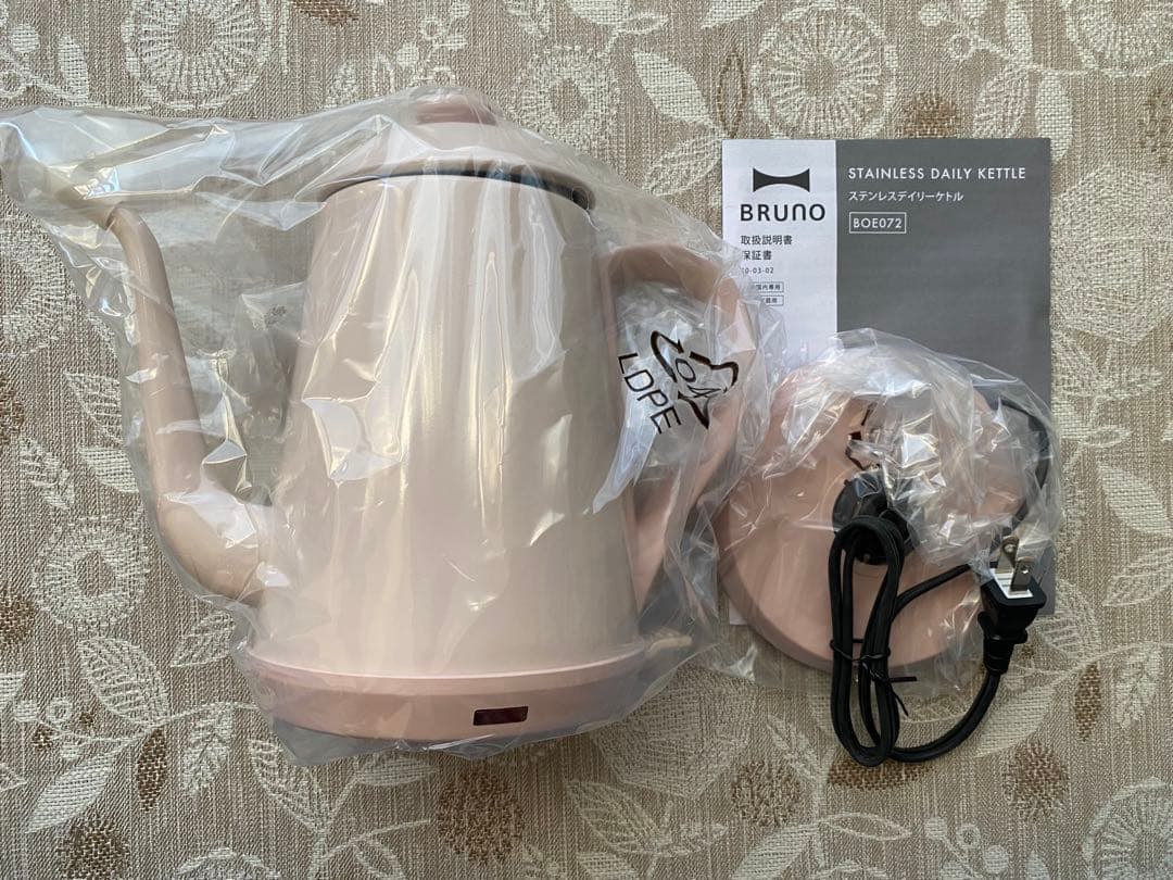 Pink-beige BRUNO stainless daily kettle wrapped in plastic with its base, power cord, and manual visible on a patterned surface.