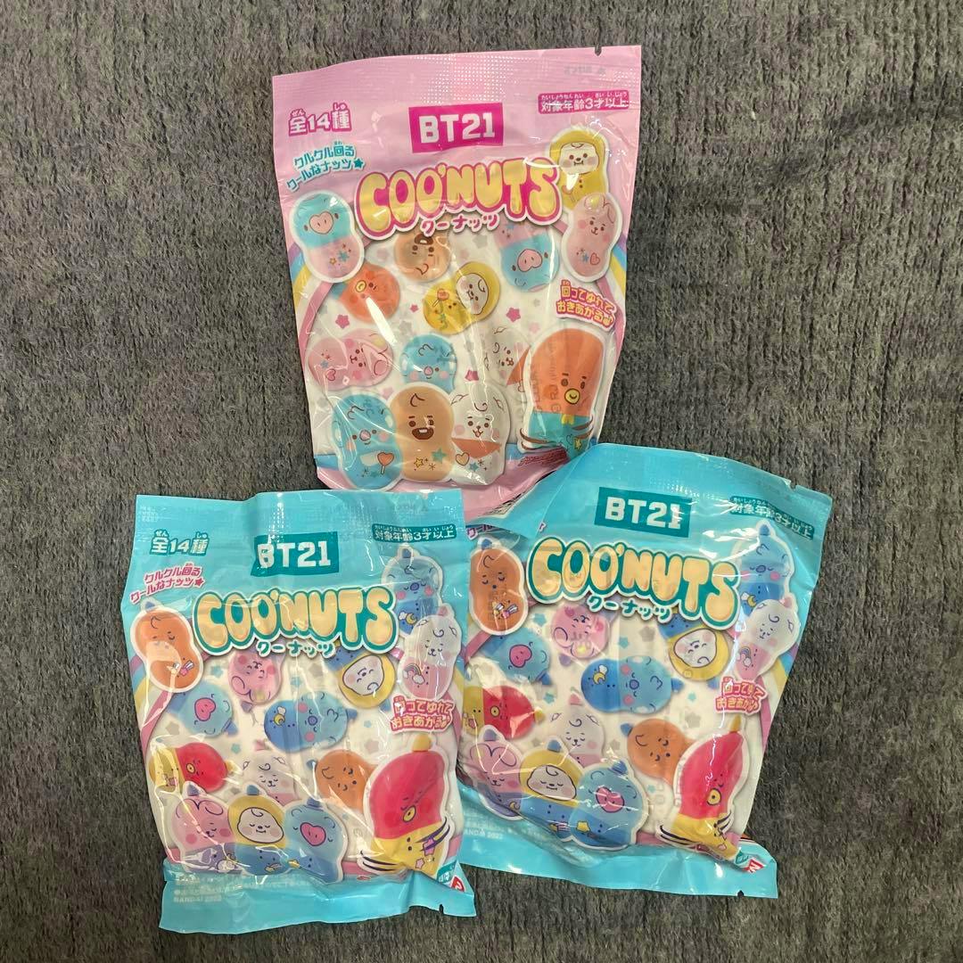 Three sealed BT21 COONUTS toy packets (one pink packet and two blue packets) displayed on a gray textured surface.