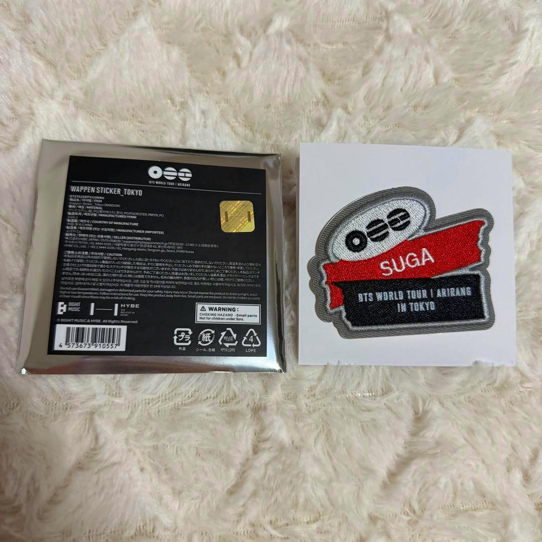A BTS ARIRANG TOKYO SUGA embroidered patch displayed on a white card next to its packaged backing on a furry surface.