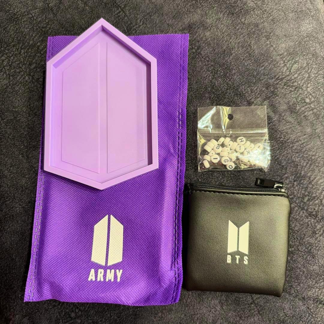 Top-down photo of a purple BTS fanclub fabric pouch with a purple hexagonal plastic plate, a small sealed bag of letter beads, and a black zip pouch printed with the BTS logo.