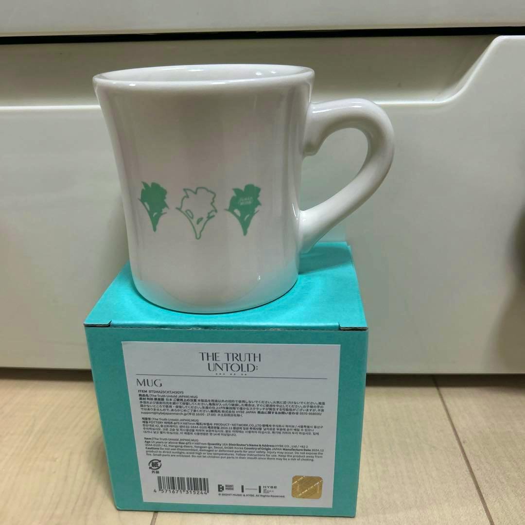 White ceramic BTS JIMIN exhibition Japan-limited mug with three green silhouette designs on its side, photographed on a teal box.