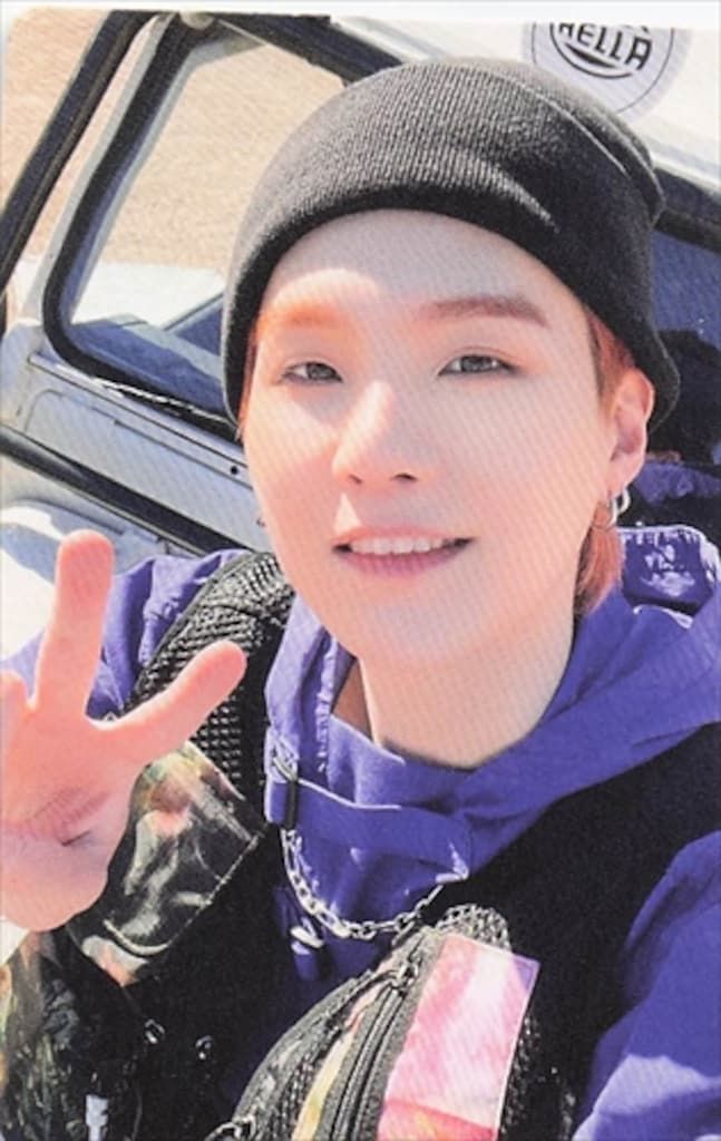 A photocard portrait of a smiling person wearing a black beanie and purple hoodie giving a peace sign.
