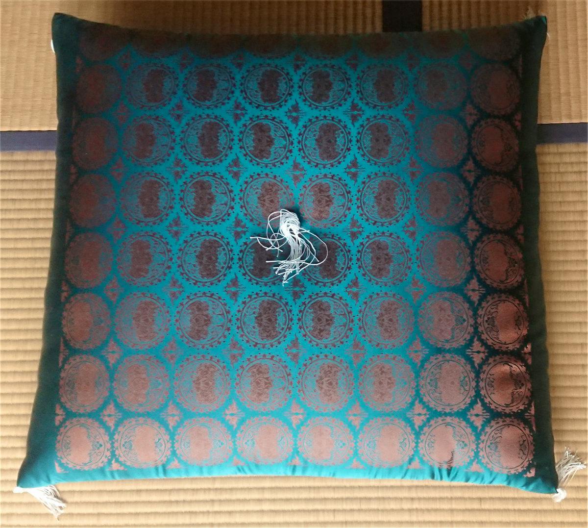 Top-down photo of a green and maroon patterned Buddhist altar cushion with white tassels placed on a tatami mat.