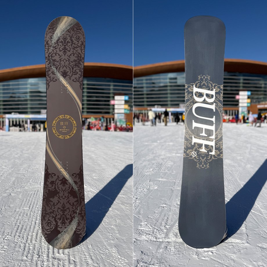 A BUFF snowboard standing upright on snow showing the top and base designs with a building in the background.