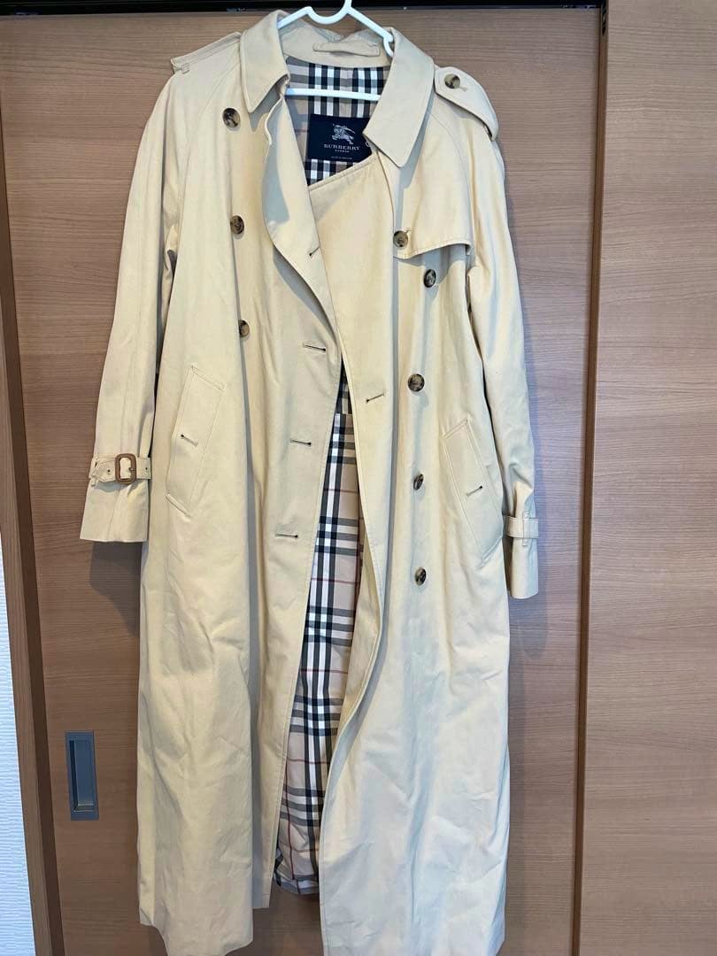 Beige Burberry men's trench coat hanging on a hanger, front view with the checked interior lining partially visible.