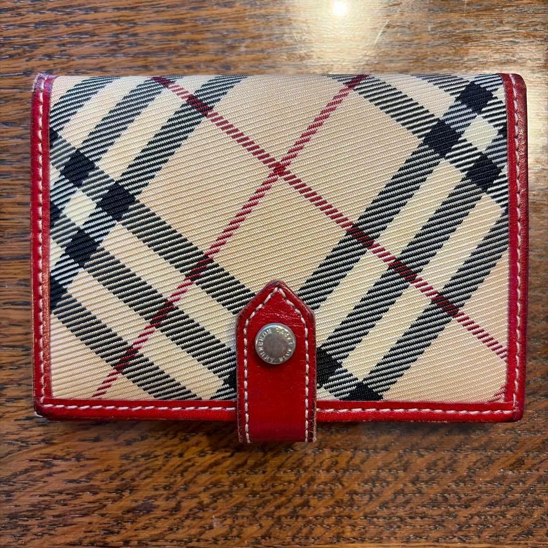 Front of a BURBERRY BLUE LABEL canvas card case with beige check pattern, red leather trim, and a snap closure.