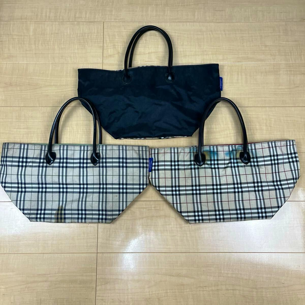 Three Burberry Blue Label tote bags (two checked canvas bags and one solid dark bag) arranged on a wooden floor.