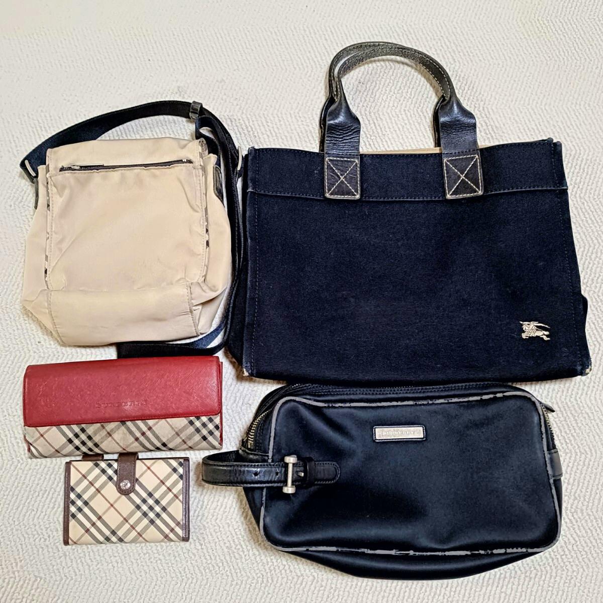 Five Burberry items laid flat: a beige shoulder bag, a black tote with leather handles, a red wallet with check-pattern trim, a small check card case, and a black zip pouch.