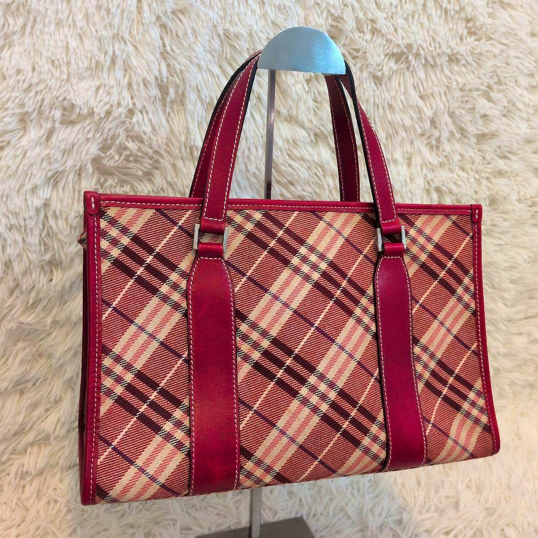 Red Burberry Blue Label Nova Check handbag with red leather trim and double handles displayed on a stand.