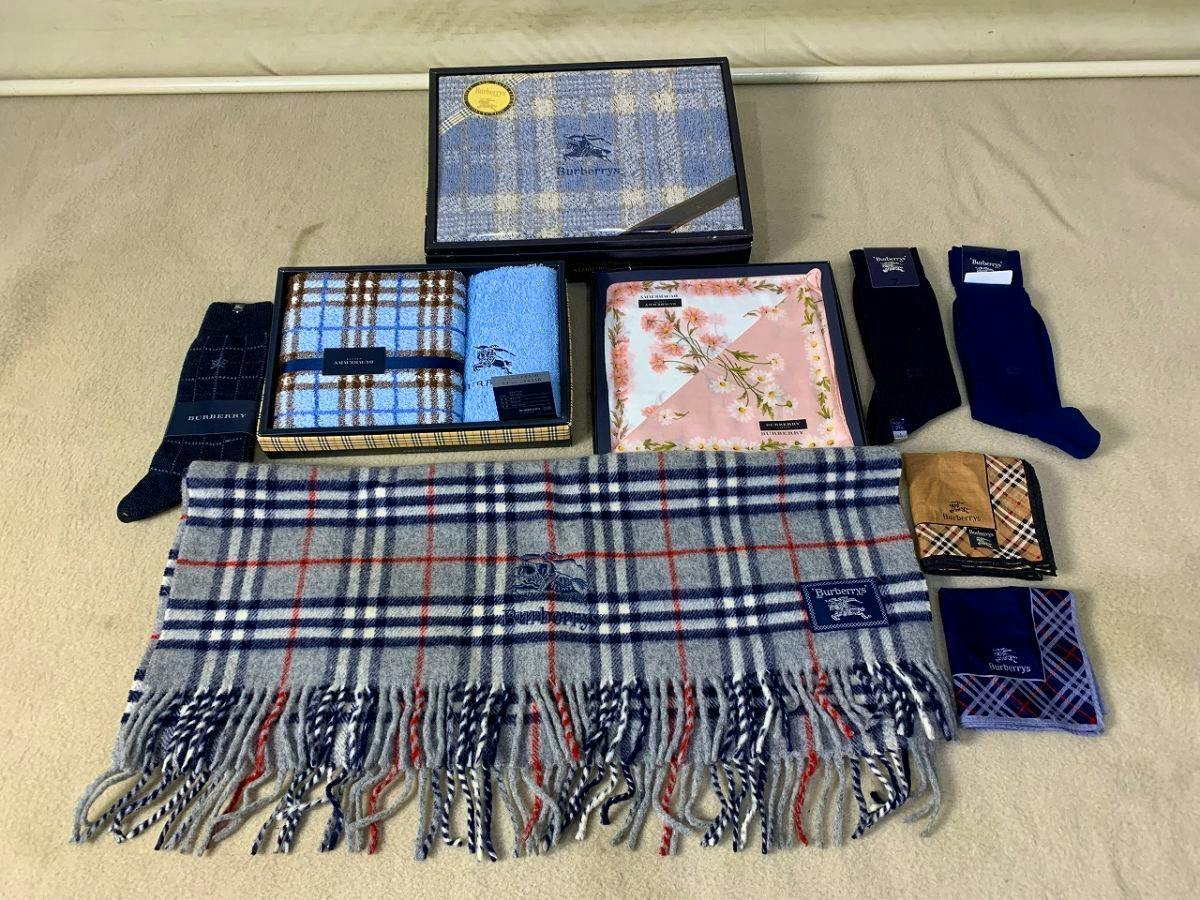 A grey wool scarf with a check tartan pattern and fringed ends laid flat with other Burberry accessories in the background.
