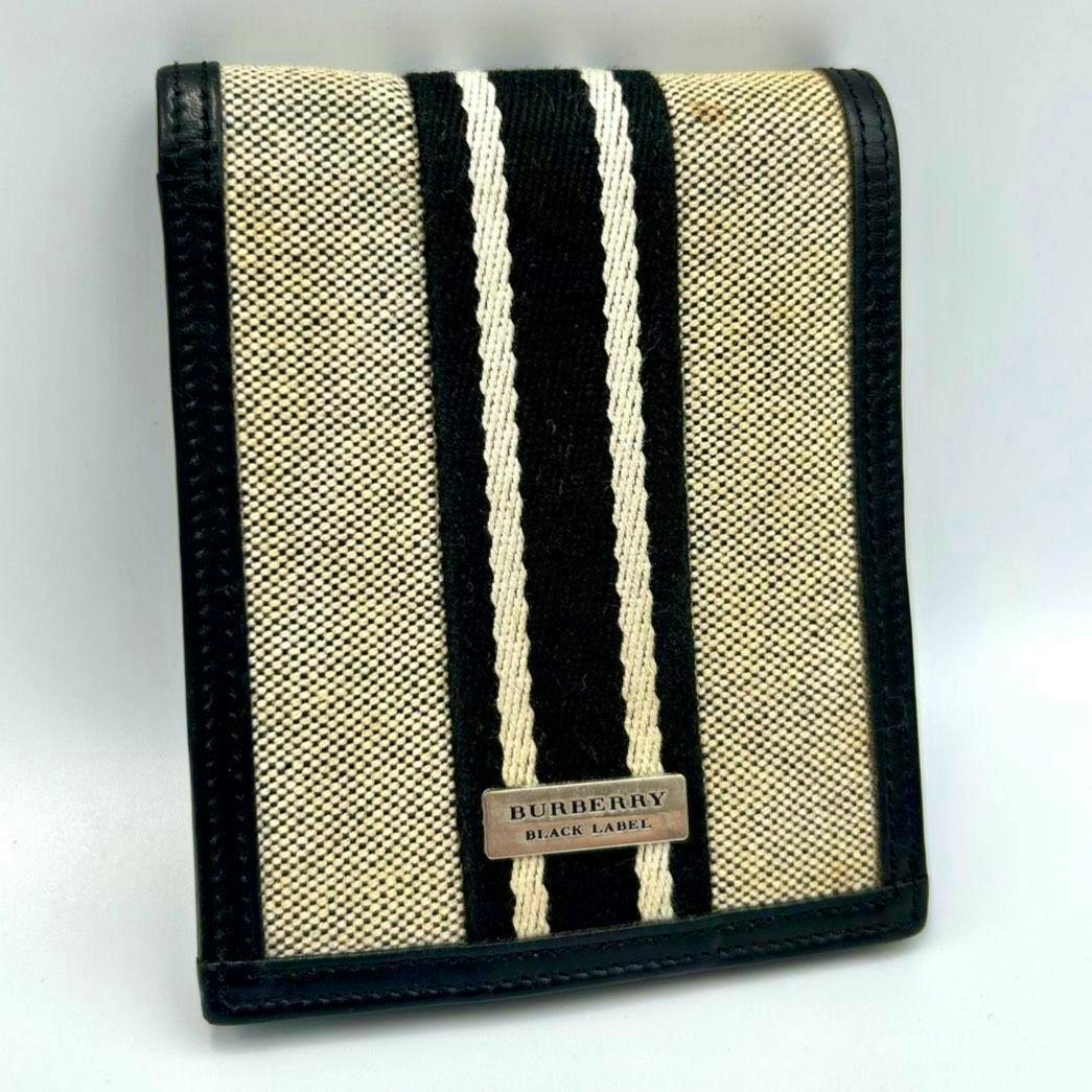 Front view of a Burberry canvas and leather bifold wallet with black and ivory center stripes and a metal "BURBERRY BLACK LABEL" plaque.