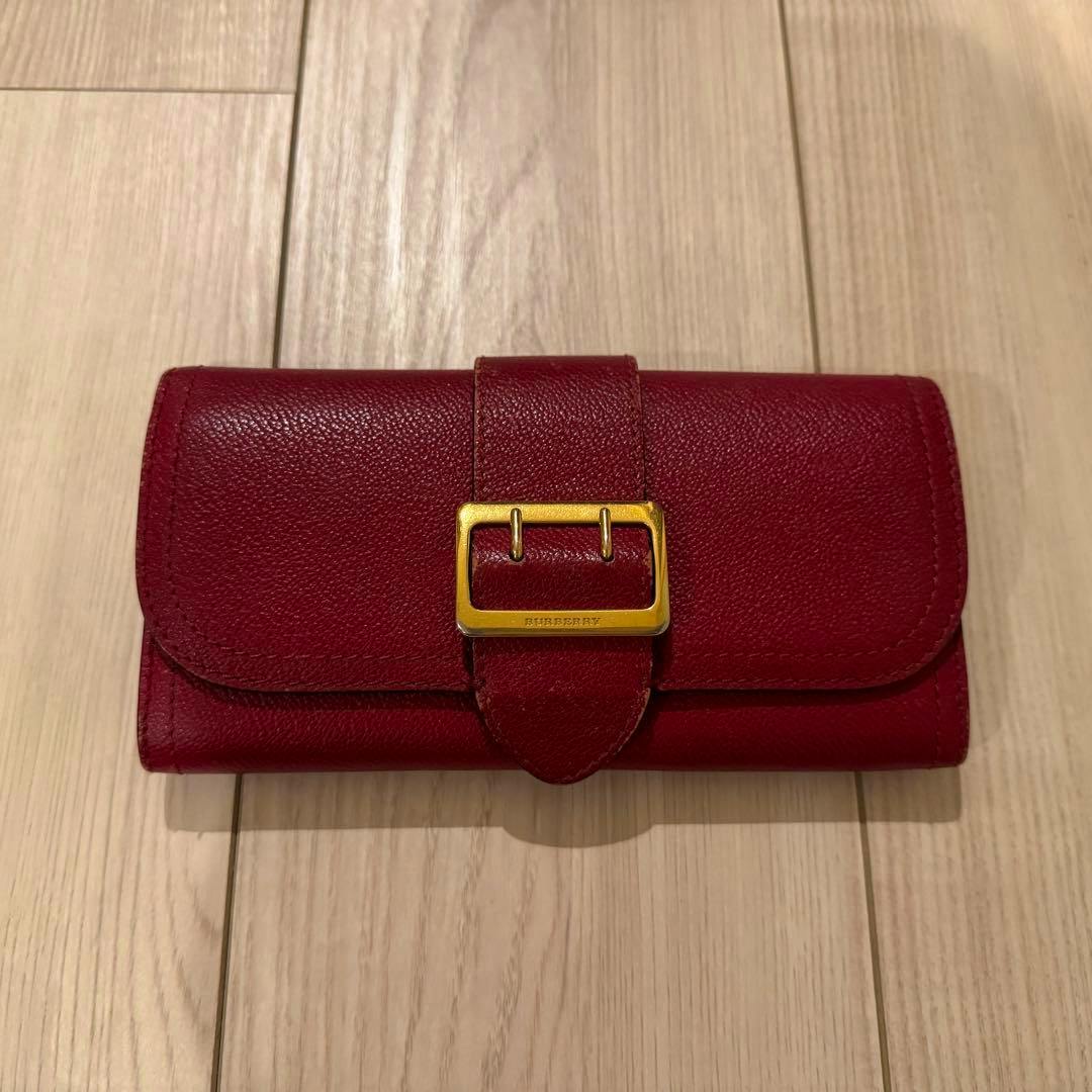 Red BURBERRY long cow leather wallet with a gold buckle closure resting on a light wooden floor.
