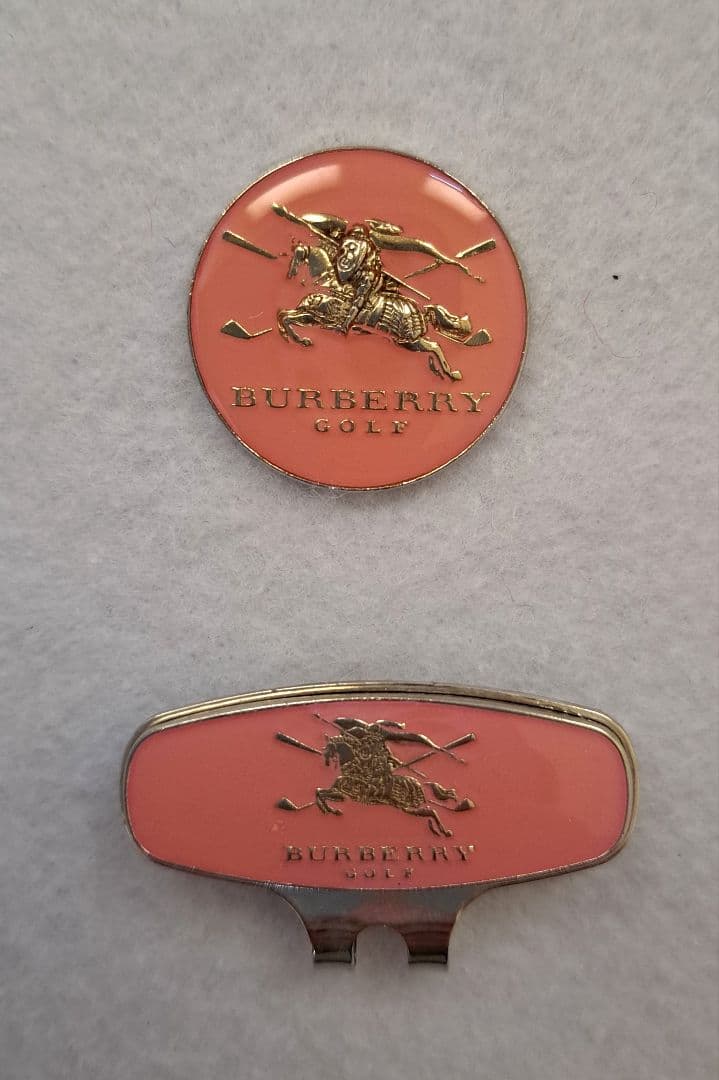 Two Burberry Golf ball markers—a round marker and a matching clip—with pink enamel and gold equestrian logo shown on a light background.