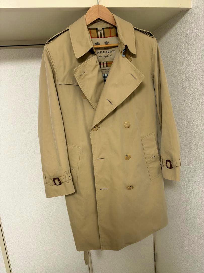 Front view of a beige Burberry men's trench coat hanging on a wooden hanger, showing the double-breasted front, pockets, and button details.