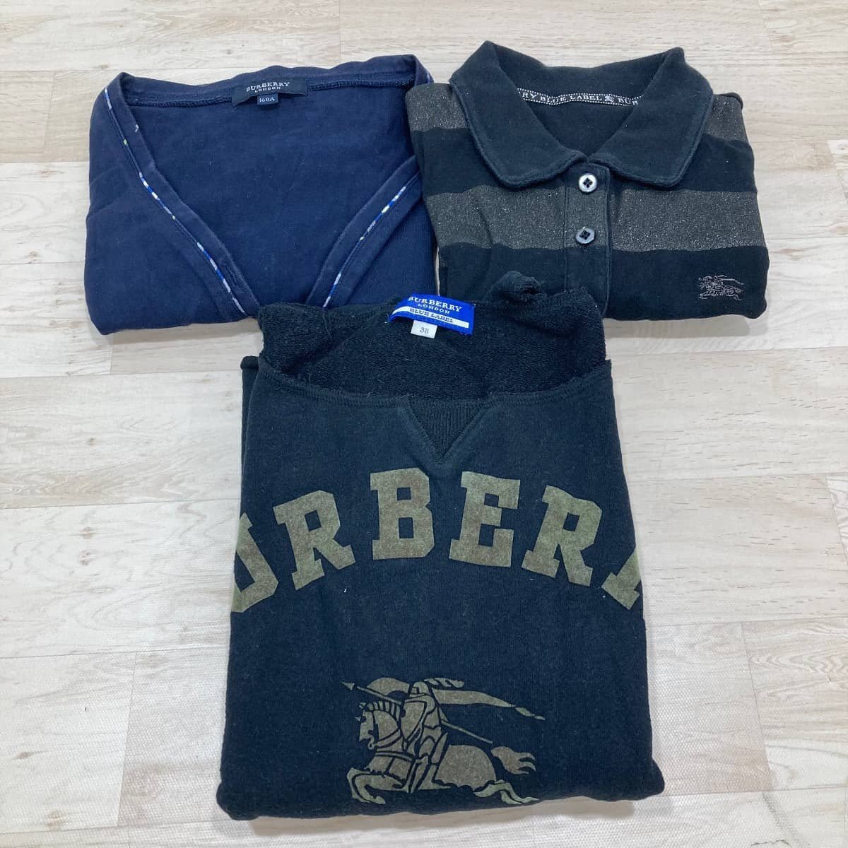 Three folded BURBERRY garments (a cardigan and two tops), one black piece with large 'BURBERRY' text and knight logo, laid on a wooden floor.
