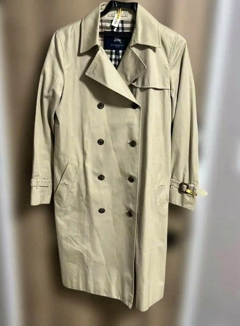 Beige men's Burberry trench coat hanging on a hanger, showing the Nova Check lining at the collar.