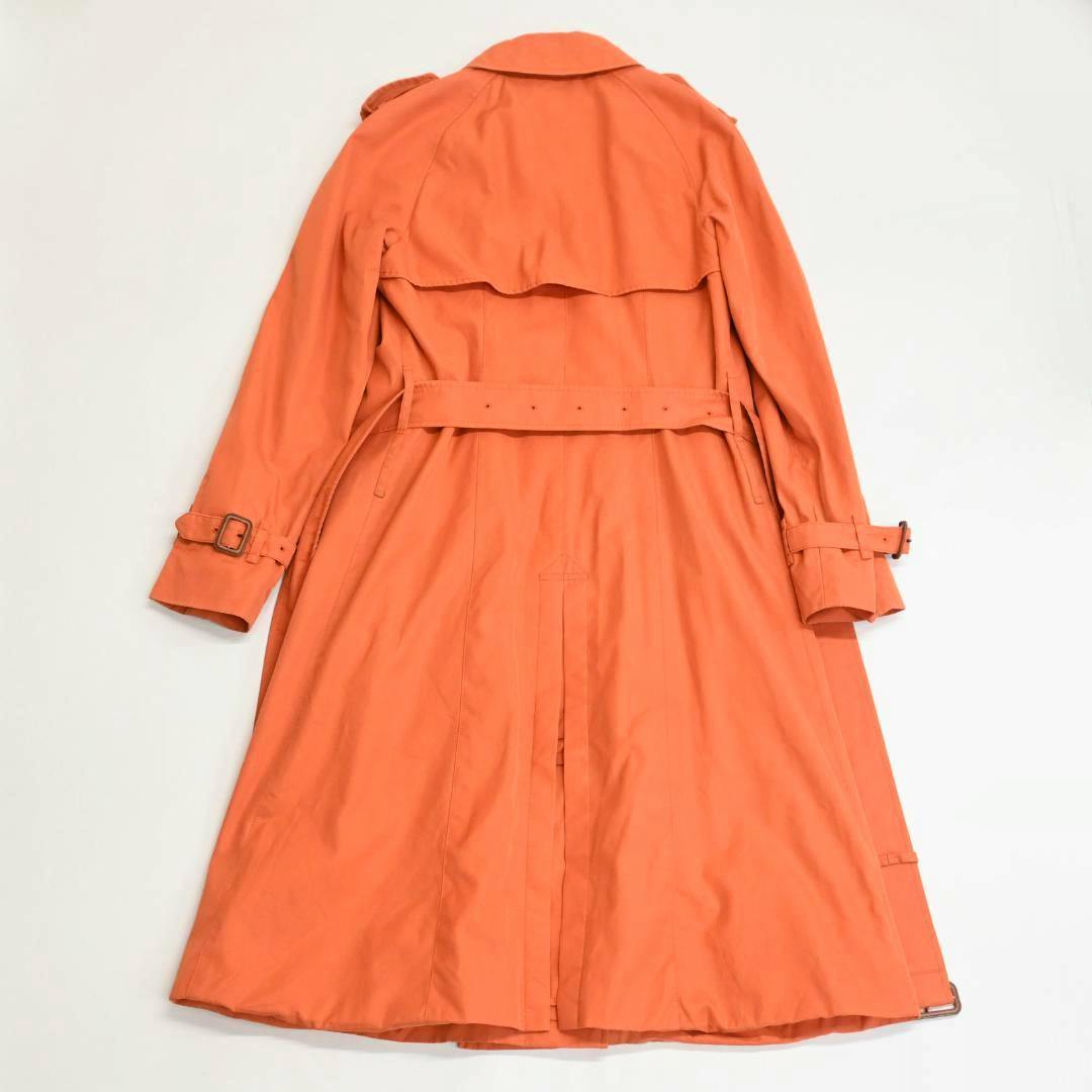 Back view of an orange women’s trench coat laid flat on a white background, showing the belt, storm flap, and sleeve buckles.