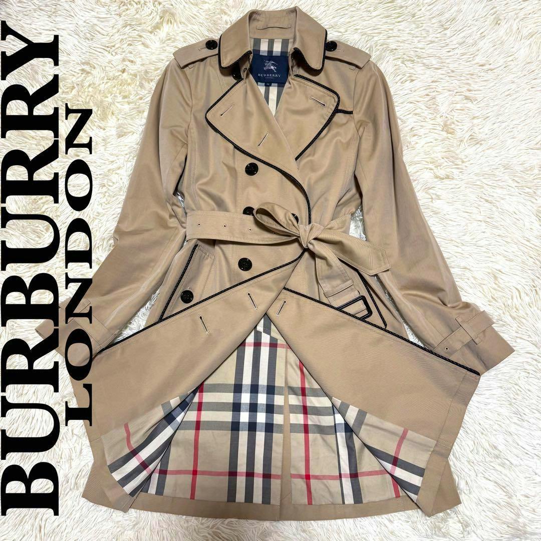 Beige Burberry trench coat with black piping, double-breasted buttons, tied belt, and checked interior lining displayed flat.