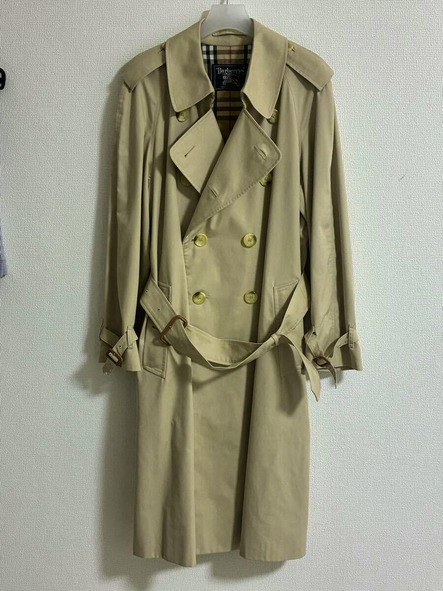 Front view of a beige double-breasted men's trench coat with a belt hanging on a hanger against a wall.