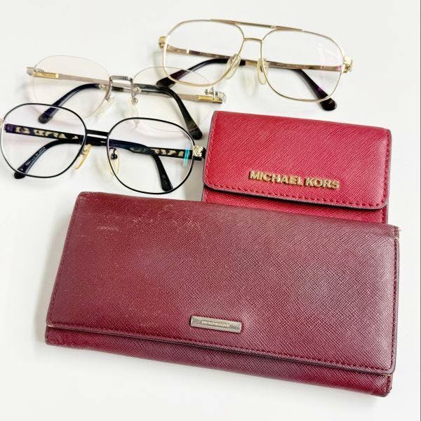 A burgundy leather wallet with a red Michael Kors card case and two pairs of eyeglasses on a white background.