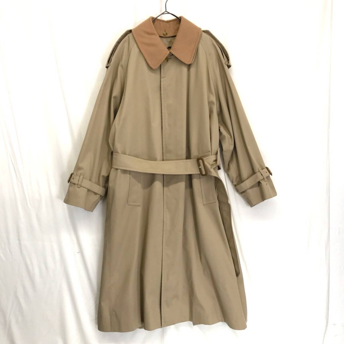 Beige Burberry trench coat with a contrasting brown collar and waist belt hung on a hanger against a white backdrop.