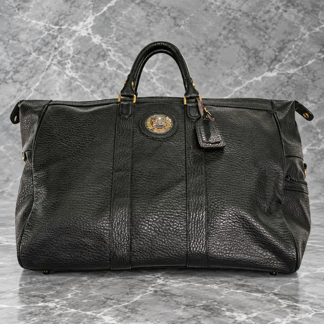 Black vintage leather Burberry Boston bag with top handles, a front emblem, and a luggage tag on a marble surface.