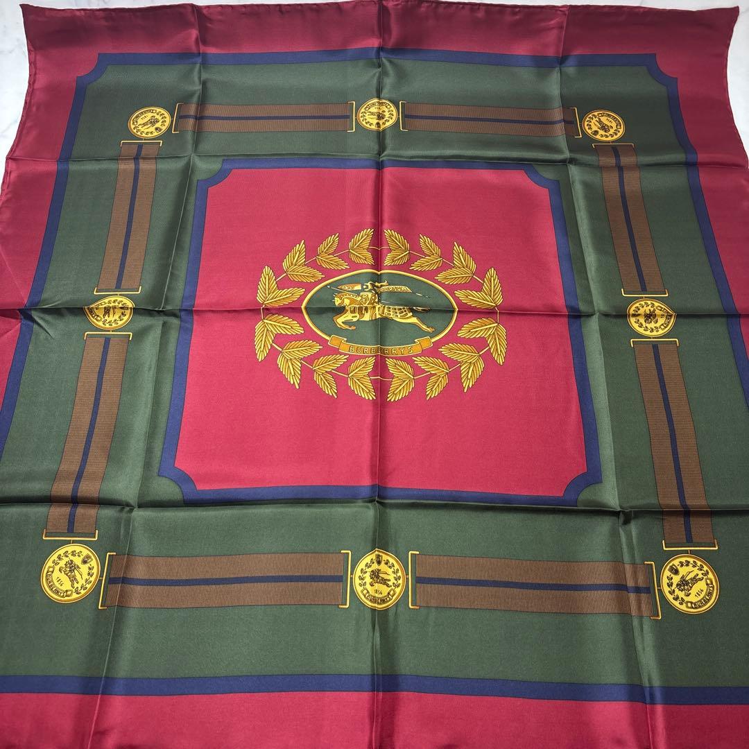 A vintage Burberry silk scarf laid flat showing a moss-green and wine-red bordered design with a central laurel emblem.