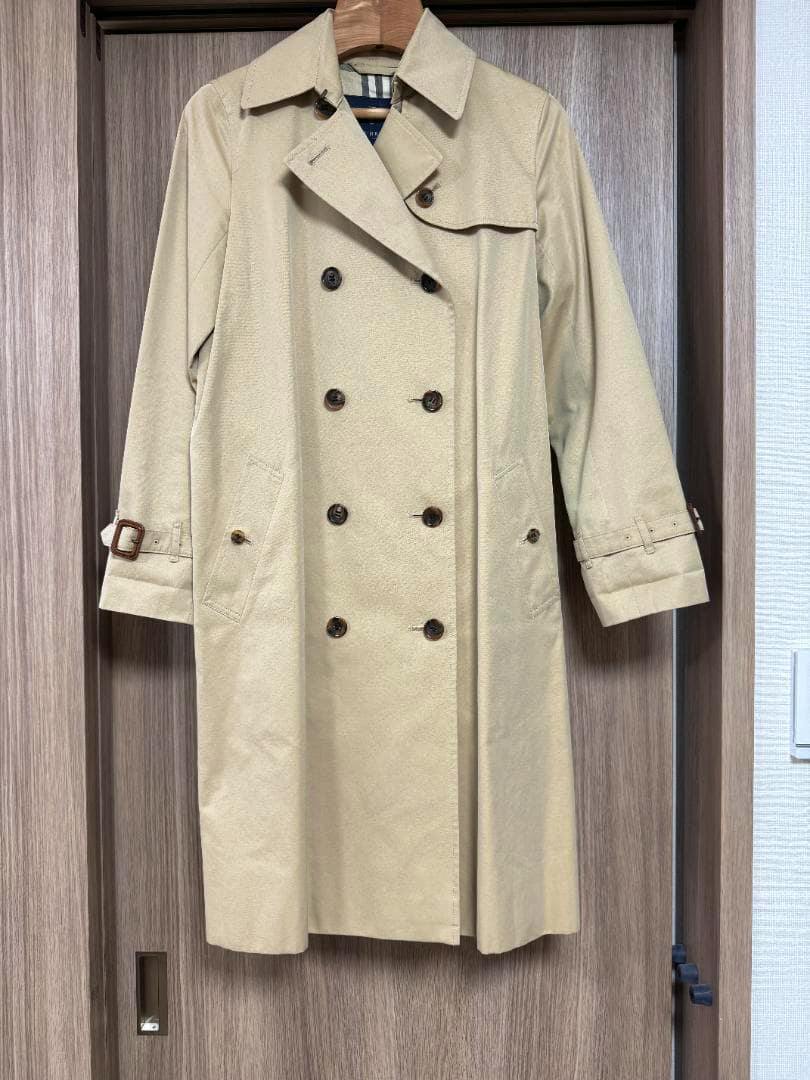 Front view of a beige women's Burberry trench coat hanging on a hanger against a wooden door.