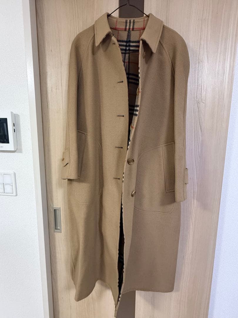 Beige BURBERRYS men's ステンカラーコート hanging on a hanger against a wooden door, shown full-length.