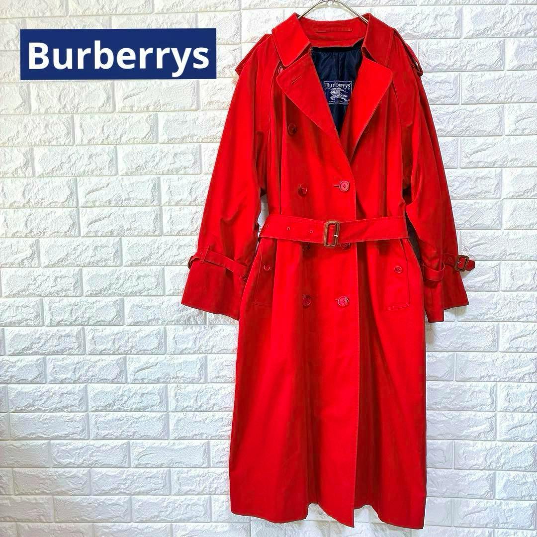 Red Burberrys women's trench coat hanging on a hanger against a white brick wall, shown full-length with belt.