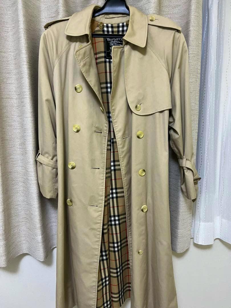 Beige Burberrys trench coat hanging on a hanger, front view showing double-breasted buttons and the checked interior lining.