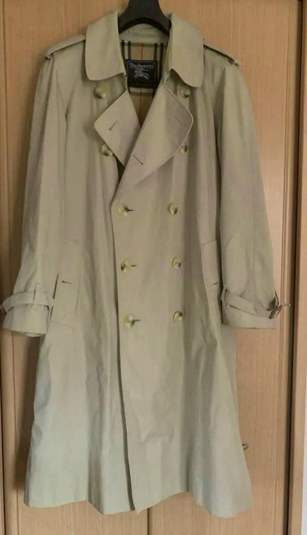 Front view of a beige men's Burberrys vintage trench coat hanging on a hanger, showing the double-breasted front and belt.