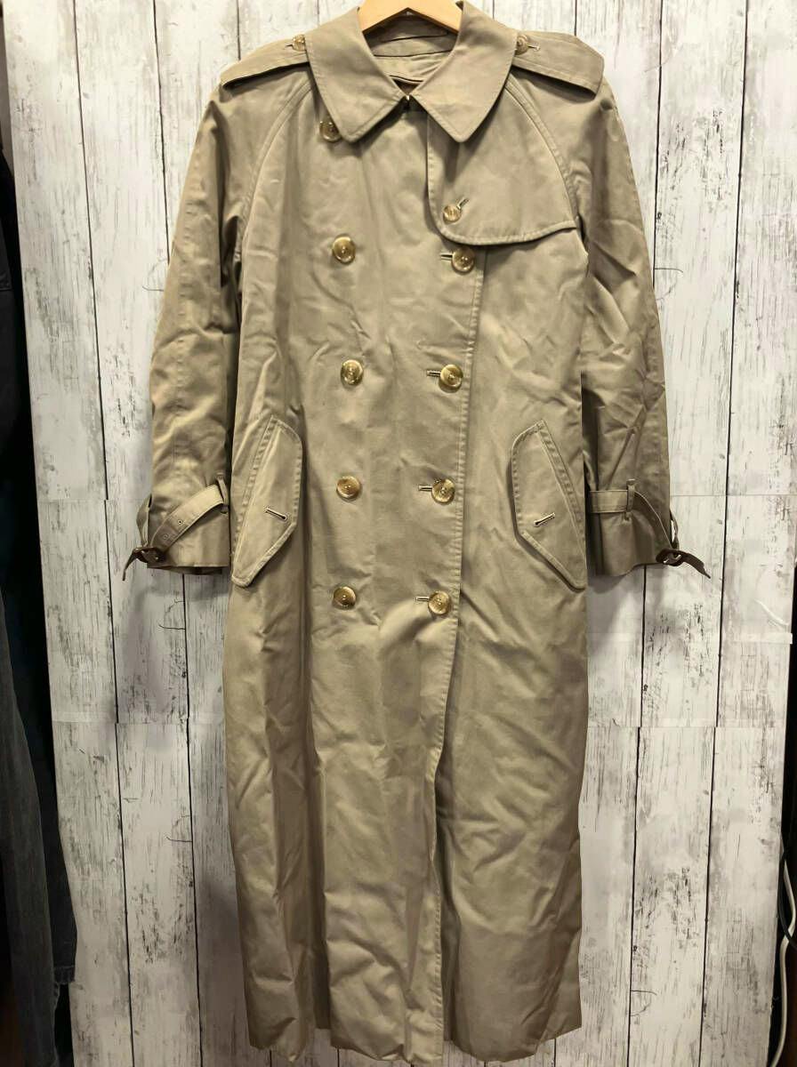 Beige Burberrys long trench coat hanging on a wooden hanger against a whitewashed wooden background.