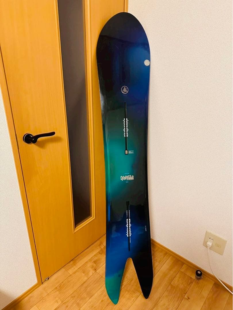 A Burton CLOUDSPLITTER snowboard with a blue-green top sheet leaning against a door on a wood floor.