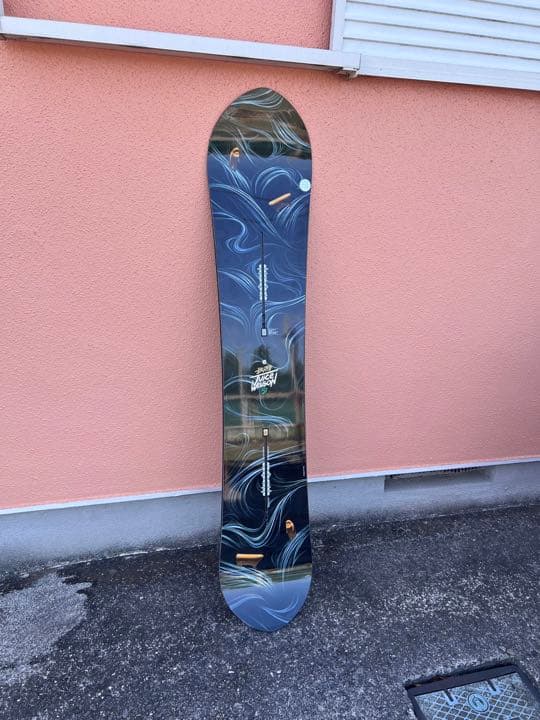 A Burton Juice Wagon 157 snowboard photographed standing upright against a pink exterior wall on pavement.