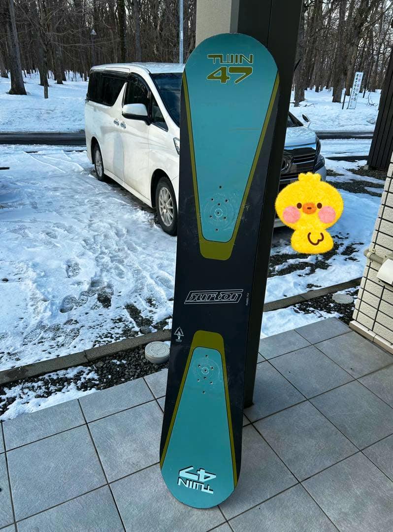 A Burton snowboard standing upright on a tiled porch with snow and a parked car visible in the background.