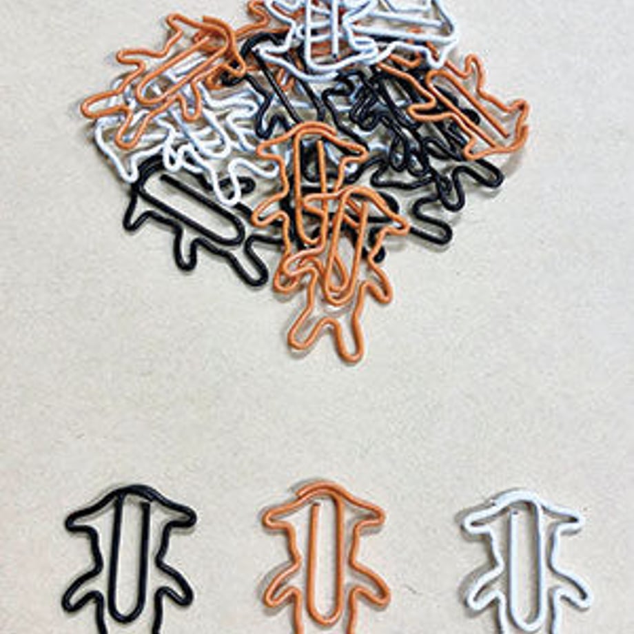 Three metal paper clips in black, orange, and white with a pile of matching clips above on a beige background.