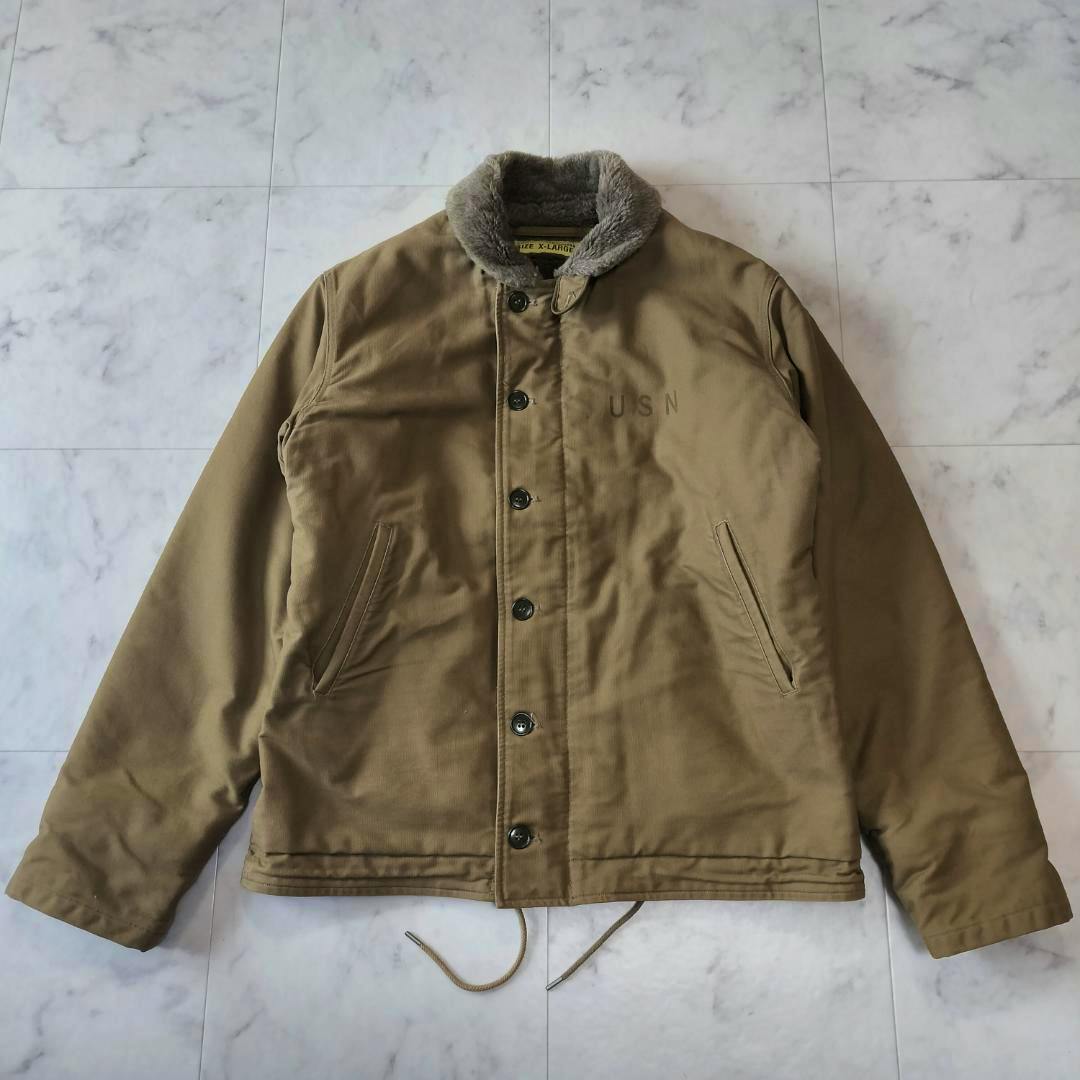Front view of a khaki Buzz Rickson's N-1 deck jacket (men's cotton) laid flat showing button closure, fur collar, pockets, drawstring, and a faint "USN" marking.