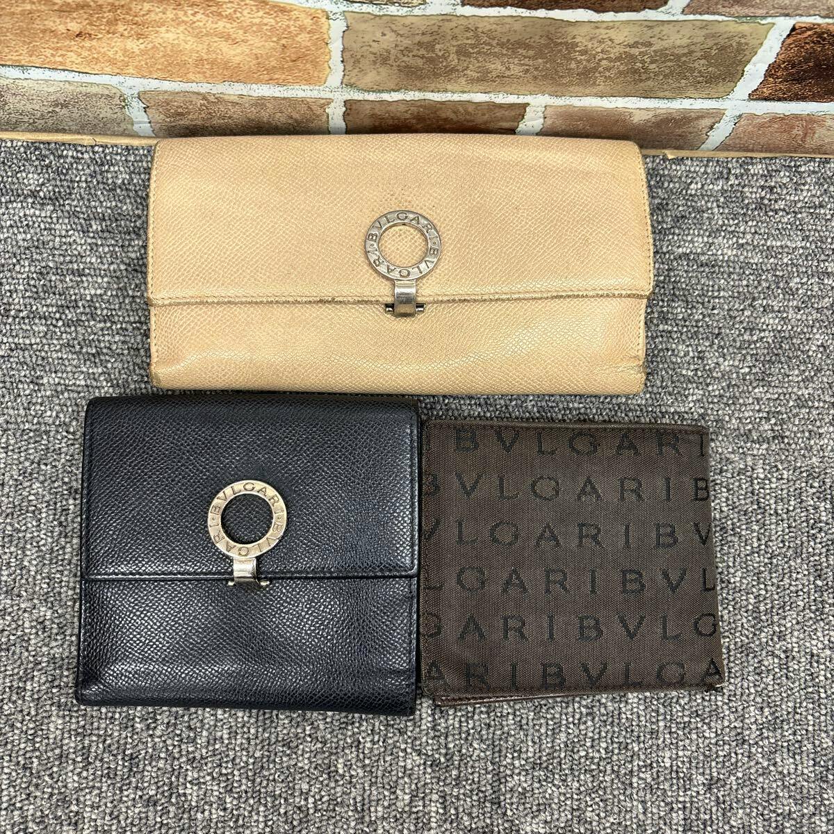 Three BVLGARI wallets — a beige long wallet, a black folded wallet with a round BVLGARI clasp, and a brown BVLGARI monogram wallet — laid on a carpeted surface.