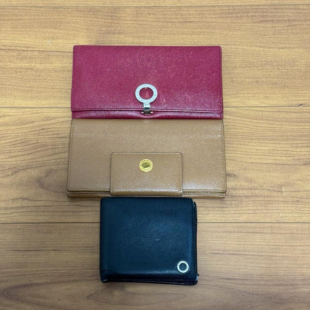 Three BVLGARI wallets displayed on a wooden surface: an open pink-and-tan long wallet and a closed black bi-fold wallet with a circular logo.