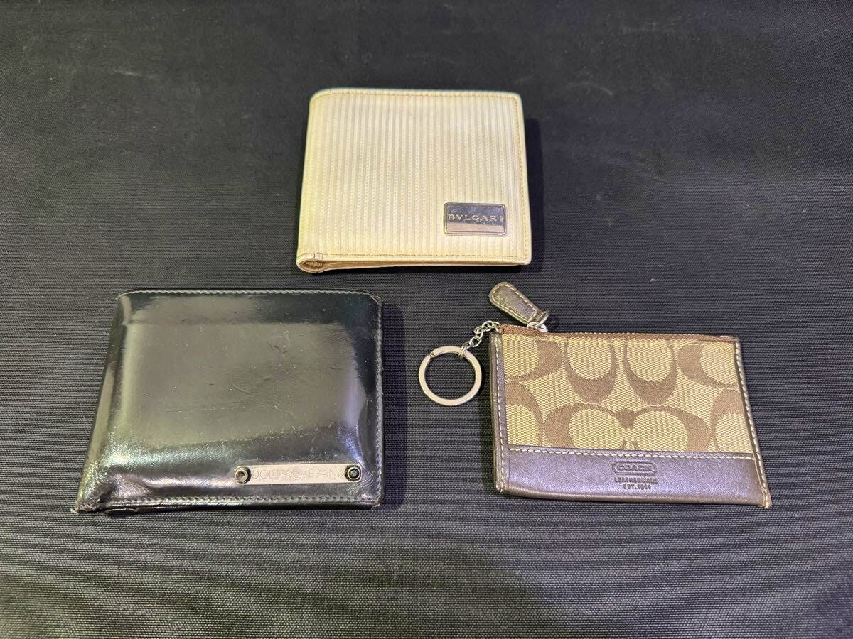 Three wallets from the bulk set—a beige BVLGARI bifold at top, a black bifold at bottom left, and a brown Coach coin case with key ring at bottom right—on a black background.