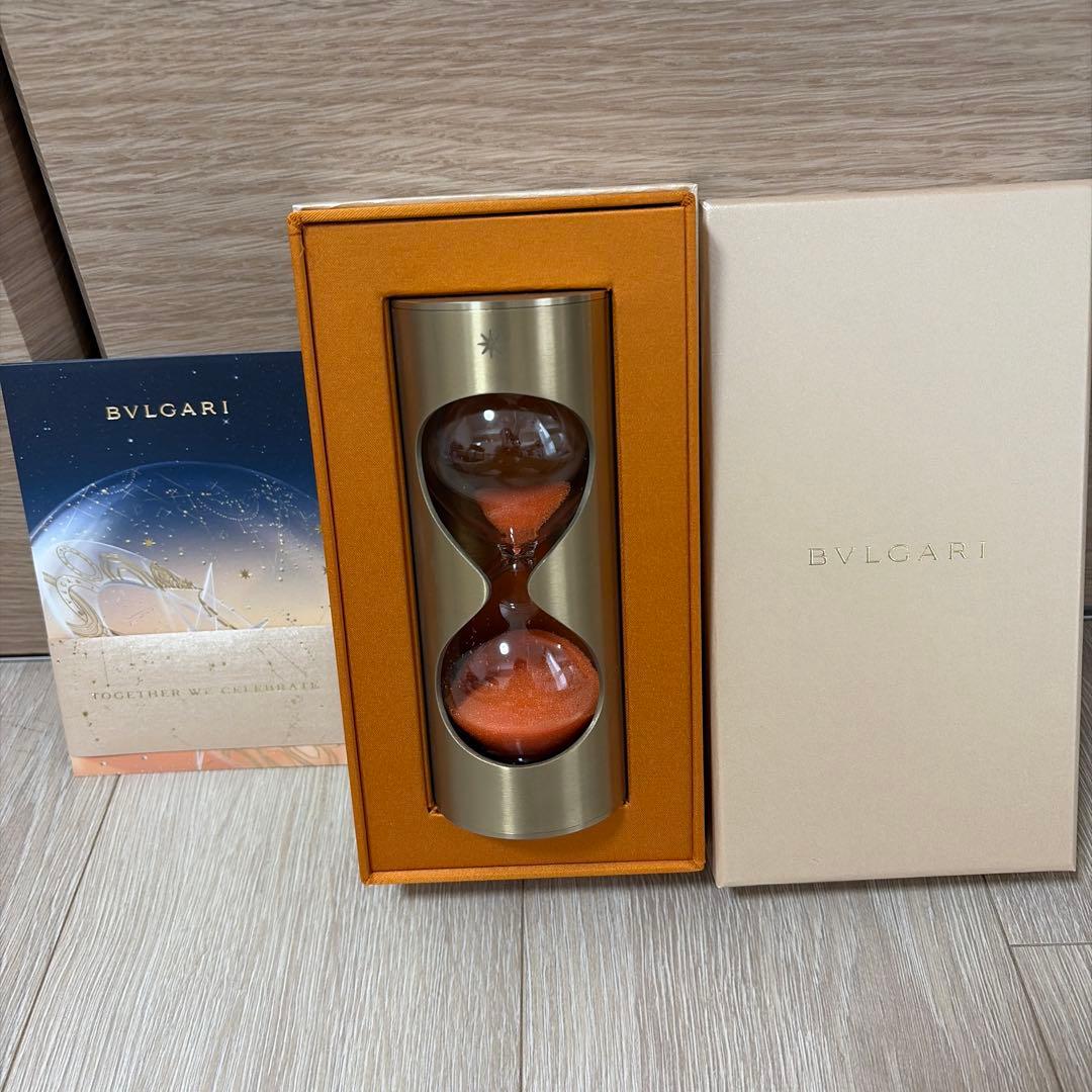BVLGARI hourglass with orange sand shown inside an orange-lined presentation box with the BVLGARI outer lid and a brochure beside it.