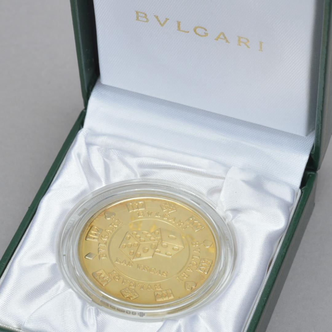 A BVLGARI casino coin in a clear plastic capsule resting on white satin inside a green presentation box with the BVLGARI logo visible on the lid.