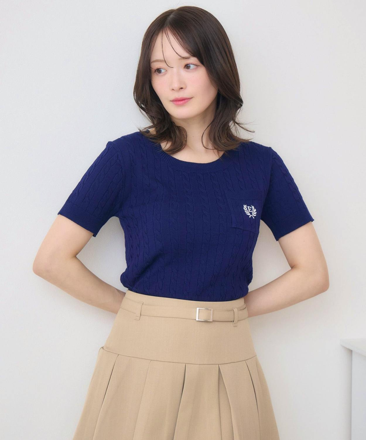 Model wearing a navy short-sleeve cable-knit women's pullover with a chest pocket and a small embroidered emblem.