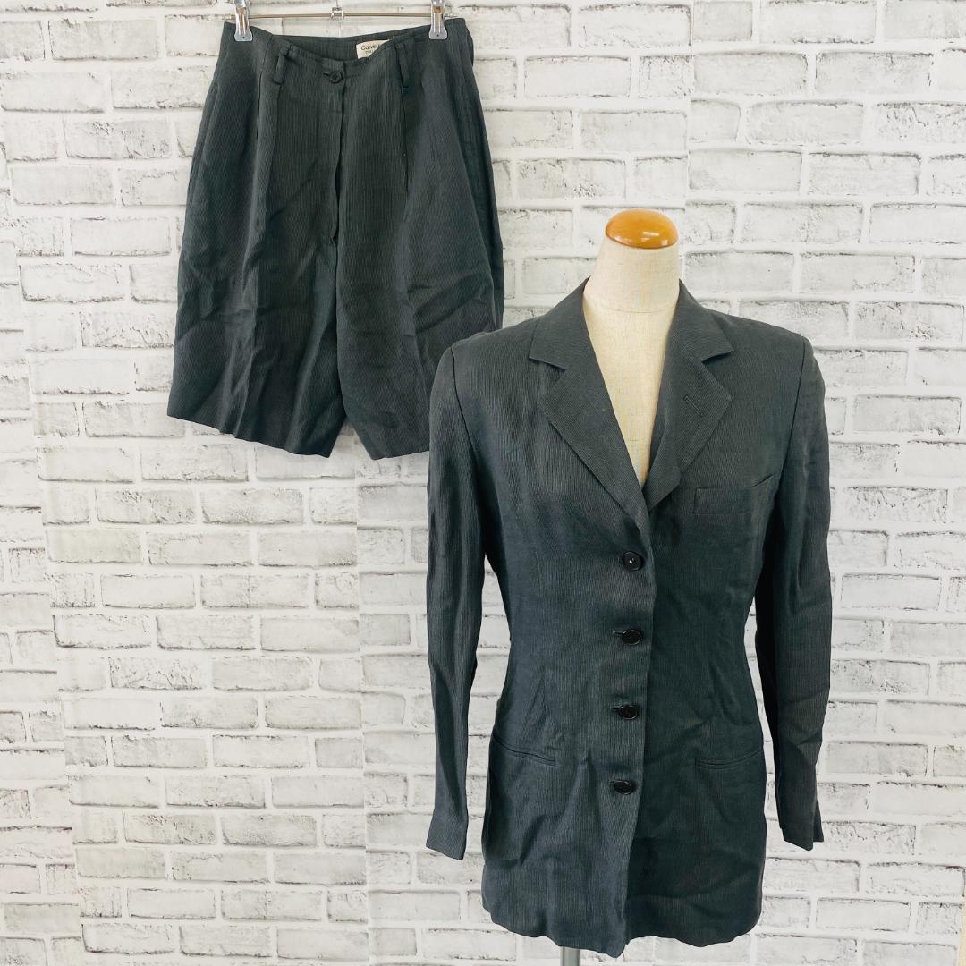Gray Calvin Klein men's suit jacket displayed on a mannequin with matching bottoms hanging behind against a white brick background.