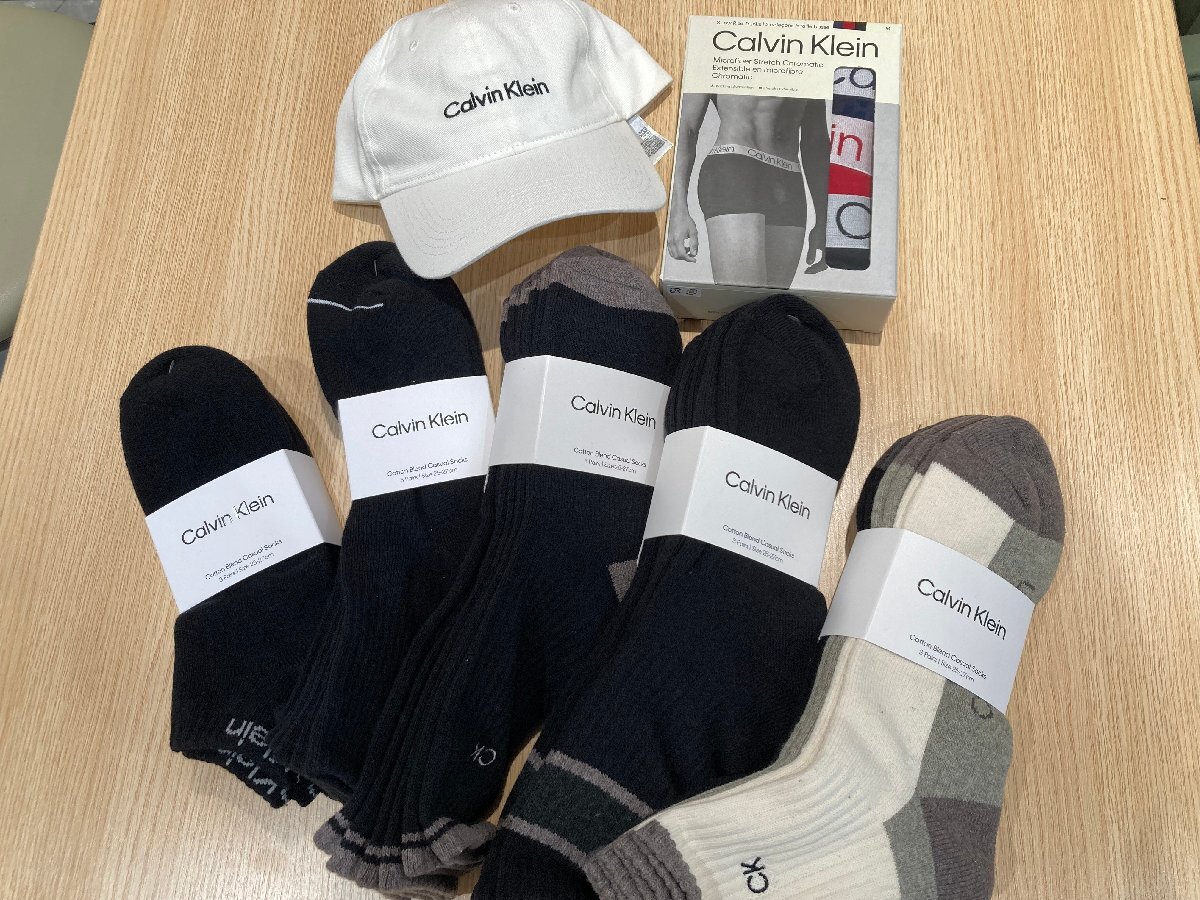 A white Calvin Klein cap, a boxed pair of Calvin Klein boxer briefs, and multiple pairs of Calvin Klein socks laid out on a wooden surface.