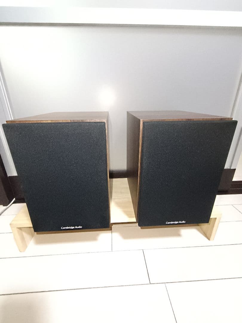 Pair of Cambridge Audio SX50 speakers with black fabric grilles placed side by side on a low wooden stand.