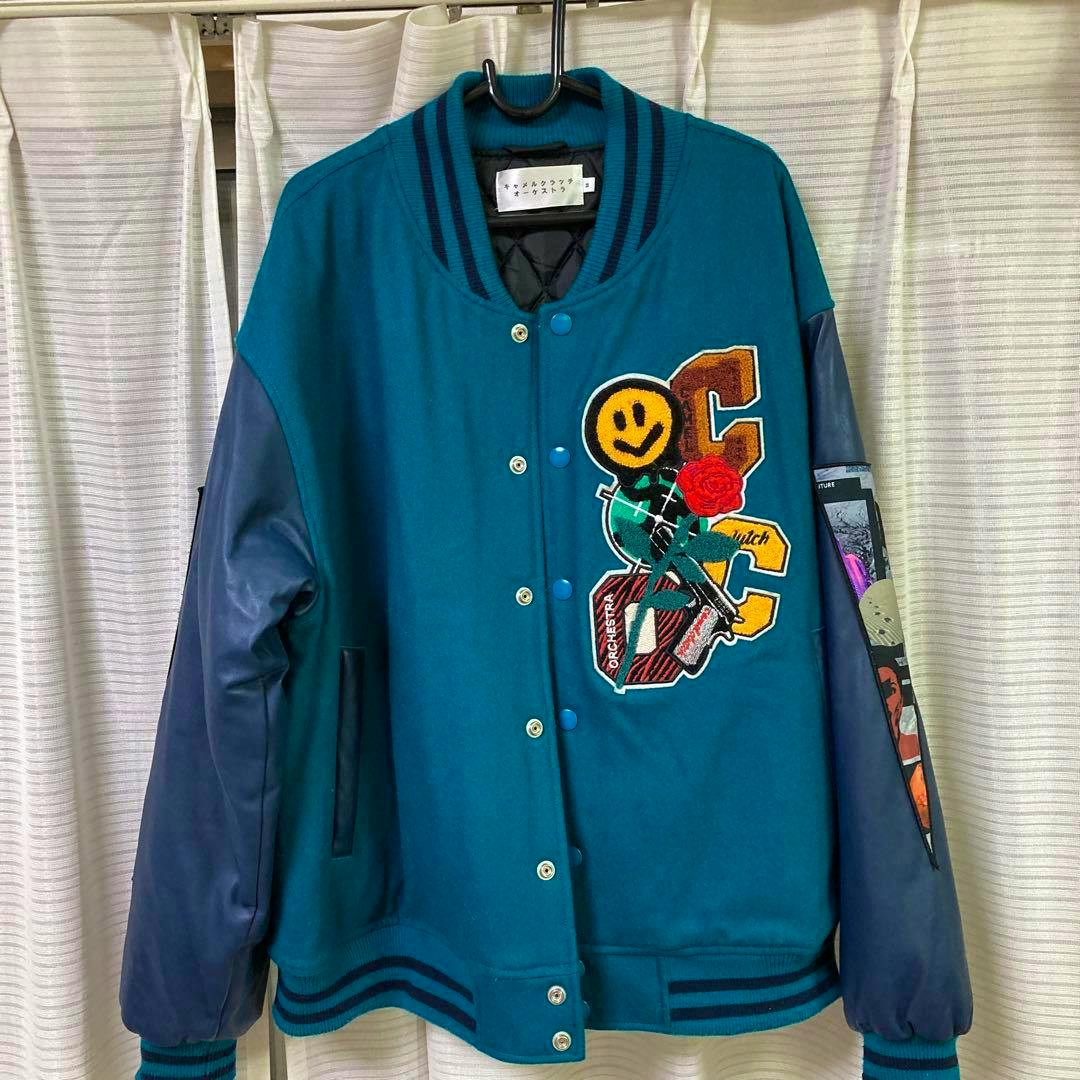Front view of the Camel Clutch Orchestra stadium jacket (スタジャン) in teal with navy sleeves and colorful embroidered patches hanging on a hanger.