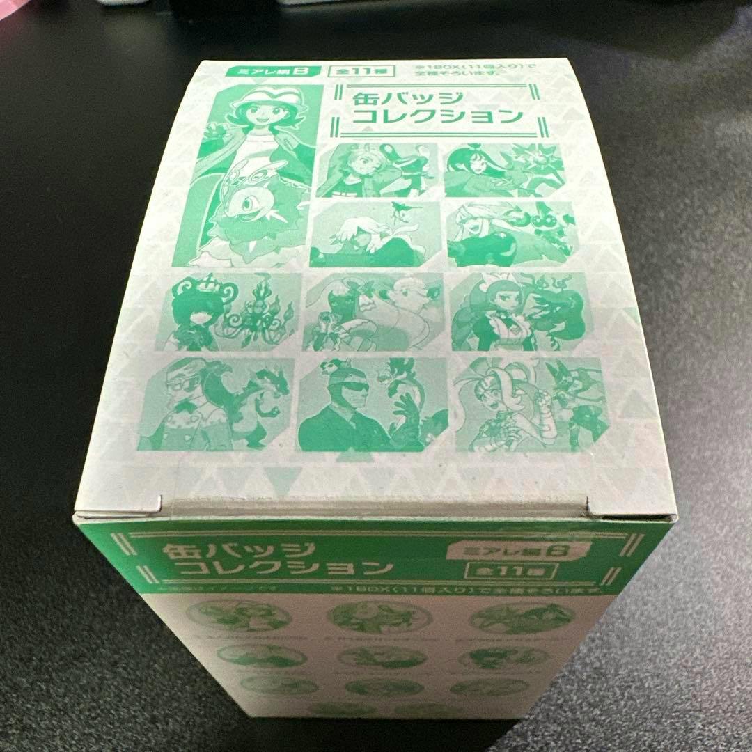 Photograph of the boxed 缶バッジコレクション〜ミアレ編 B〜 badge collection box showing printed illustrations of the included badges on the packaging.