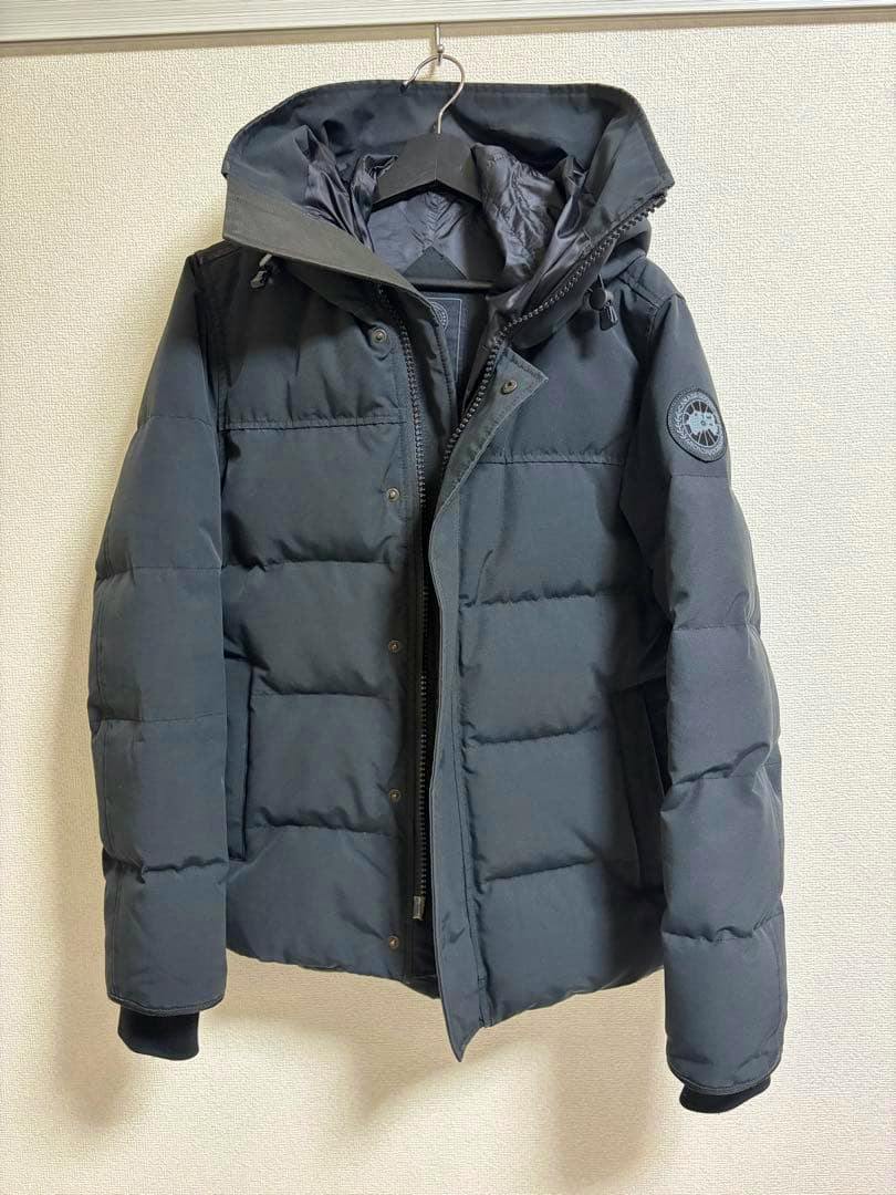 Black CANADA GOOSE Macmillan parka (men's) hanging on a hanger showing the front view with hood and sleeve logo.